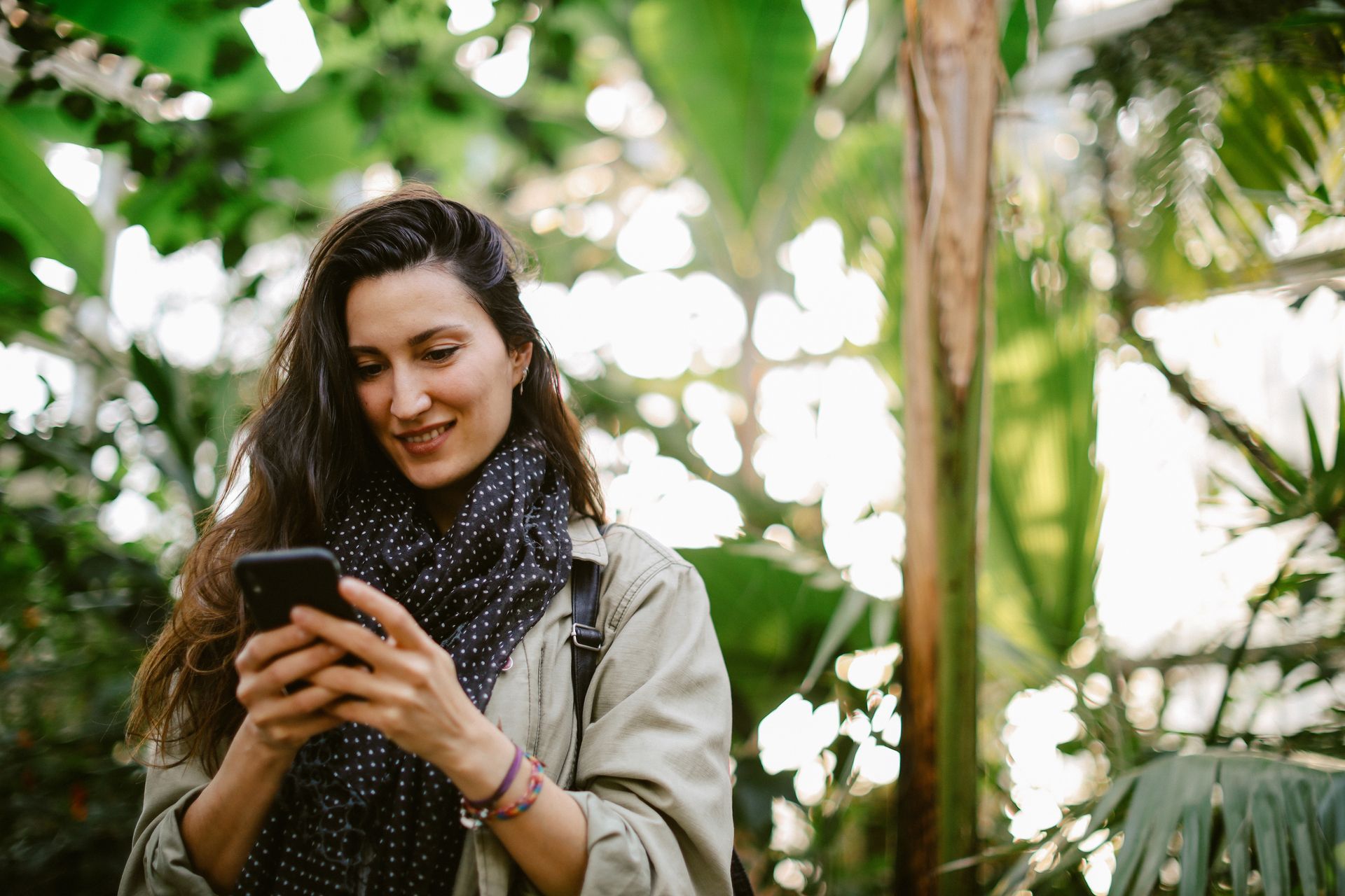 Žena venku se usmívá do mobilního telefonu, obklopená stromy.